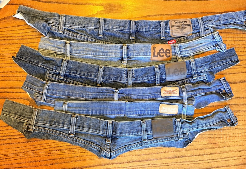 May include: An assortment of denim jean waistbands in various shades of blue. The waistbands are from different brands, including Wrangler, Lee, and Levi's, each with its brand label. The jeans are laid out on a wooden surface.