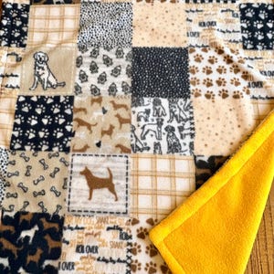 May include: A patchwork dog blanket with various patterns, including paw prints, dog silhouettes, and bone shapes. The blanket features squares in shades of brown, beige, black, and white, with a bright yellow border.