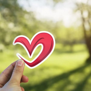 May include: A red heart-shaped sticker with a white outline, held in a hand against a blurred green background.