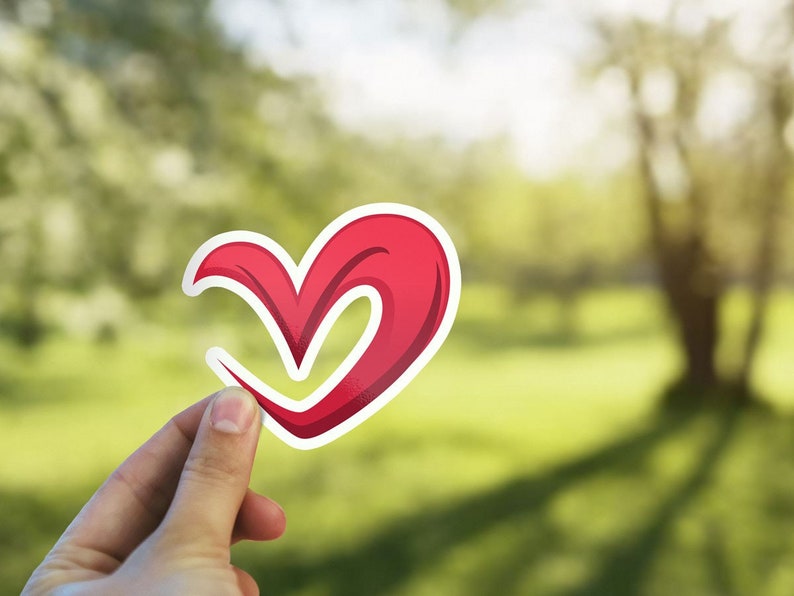 May include: A red heart-shaped sticker with a white outline, held in a hand against a blurred green background.