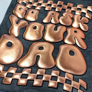 May include: A dark grey t-shirt with a rose gold foiled design that reads "Go Ask Your Dad" in a bubble font. The design is outlined in black and has a checkered pattern above and below the text.
