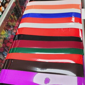 Op de afbeelding: Een selectie van vinylplaten in verschillende kleuren, waaronder zwart, paars, bruin, rood, groen, donkerrood, wit en blauw. De platen zijn op elkaar gestapeld en hebben een glanzende afwerking.