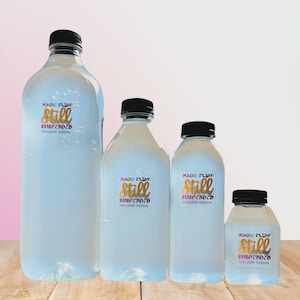 Puede incluir: Cuatro botellas de plástico transparente de diferentes tamaños con tapas negras están dispuestas sobre una superficie de madera. Cada botella tiene una etiqueta con el texto "MAGIC PLAV Still SUSPENDED Monglobe Solution" en oro y rojo.