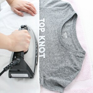 Puede incluir: Una persona planchando una camiseta gris con las palabras "TOP KNOT" impresas en la parte delantera. La camiseta est&aacute; sobre una superficie blanca y se puede ver una toalla rosa en el fondo.