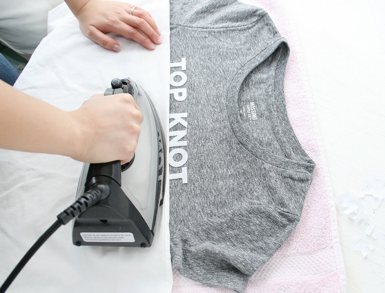Puede incluir: Una persona planchando una camiseta gris con las palabras "TOP KNOT" impresas en la parte delantera. La camiseta est&aacute; sobre una superficie blanca y se puede ver una toalla rosa en el fondo.