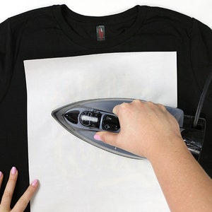 May include: A person ironing a black t-shirt with a white piece of paper on top of it.