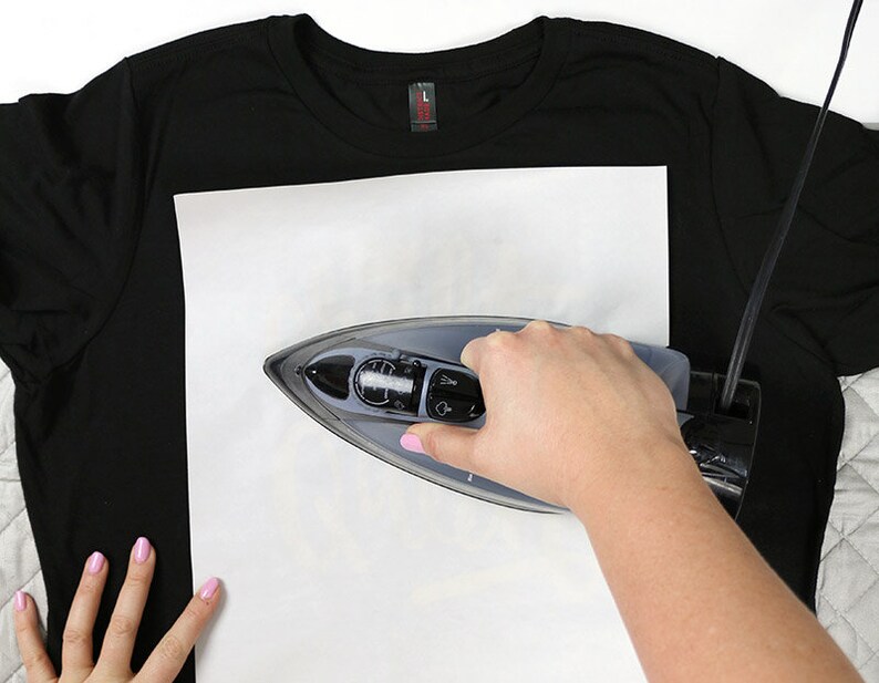 May include: A person ironing a black t-shirt with a white piece of paper on top of it.