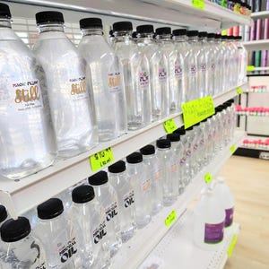 May include: Clear plastic bottles with black caps are displayed on white shelves. Some bottles have labels that read "Magic Flow Still" in gold lettering. Other bottles are labeled "Magic Flow Mineral Oil".