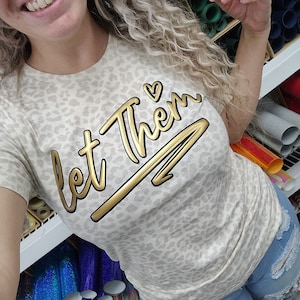 May include: A beige t-shirt with a leopard print pattern and gold lettering that says "Let Them" with a heart symbol. The shirt is being worn by a person.