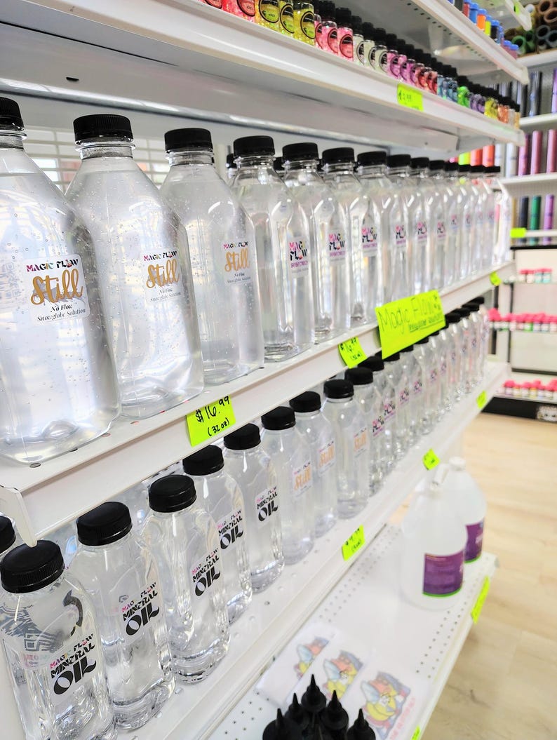 May include: Clear plastic bottles with black caps are displayed on white shelves. Some bottles have labels that read "Magic Flow Still" in gold lettering. Other bottles are labeled "Magic Flow Mineral Oil".