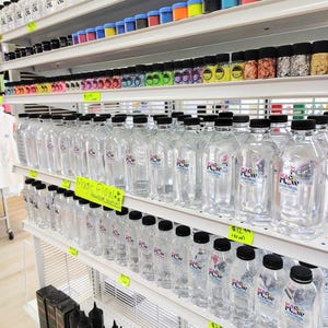 May include: Shelves stocked with various craft supplies, including clear plastic bottles with black caps labeled "Magic Flow." Other items include small jars of glitter, colorful powders, and bottles of oil. The store is well-lit and organized.
