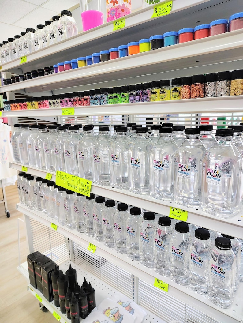 May include: Shelves stocked with various craft supplies, including clear plastic bottles with black caps labeled "Magic Flow." Other items include small jars of glitter, colorful powders, and bottles of oil. The store is well-lit and organized.