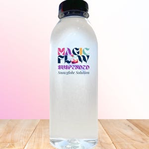 May include: A clear plastic bottle with a black cap, filled with a liquid. The bottle has a colorful label that reads "MAGIC FLOW SUSPENDED Snowglobe Solution". The bottle is sitting on a wooden surface.