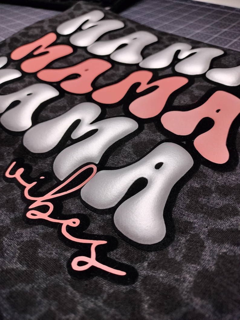 May include: Black and white leopard print fabric with the words "Mama" in silver and pink lettering. The word "Mama" is repeated three times, with the last "Mama" being in pink. The words "& babes" are written in pink below the word "Mama".
