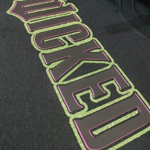 Może przedstawiać: Czarny T-shirt z napisem "WICKED" w dużych, błyszczących zielonych i r&oacute;żowych literach.