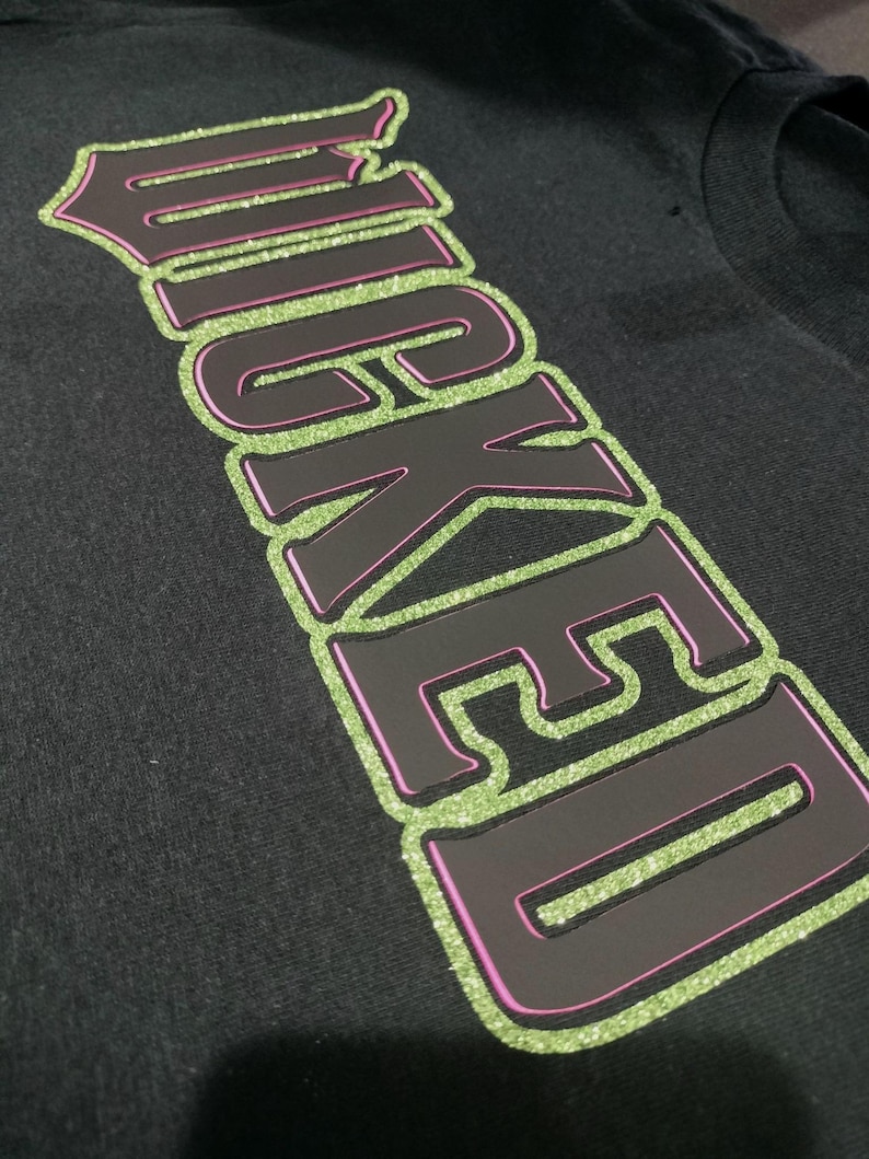 Może przedstawiać: Czarny T-shirt z napisem "WICKED" w dużych, błyszczących zielonych i r&oacute;żowych literach.