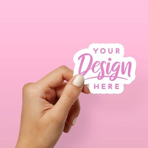 May include: A white sticker with rounded edges and a pink design that says "YOUR Design HERE". The sticker is being held by a hand against a pink background.