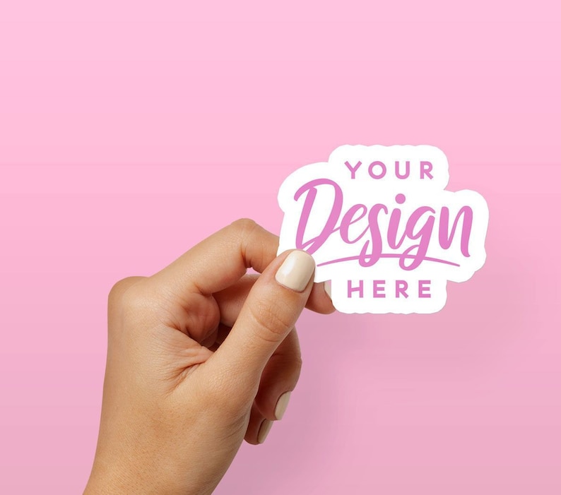 May include: A white sticker with rounded edges and a pink design that says "YOUR Design HERE". The sticker is being held by a hand against a pink background.