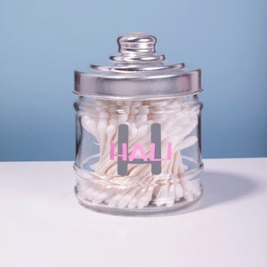 May include: A clear glass jar with a silver lid filled with cotton swabs. The jar has a gray label with the word "HALI" written in pink letters.
