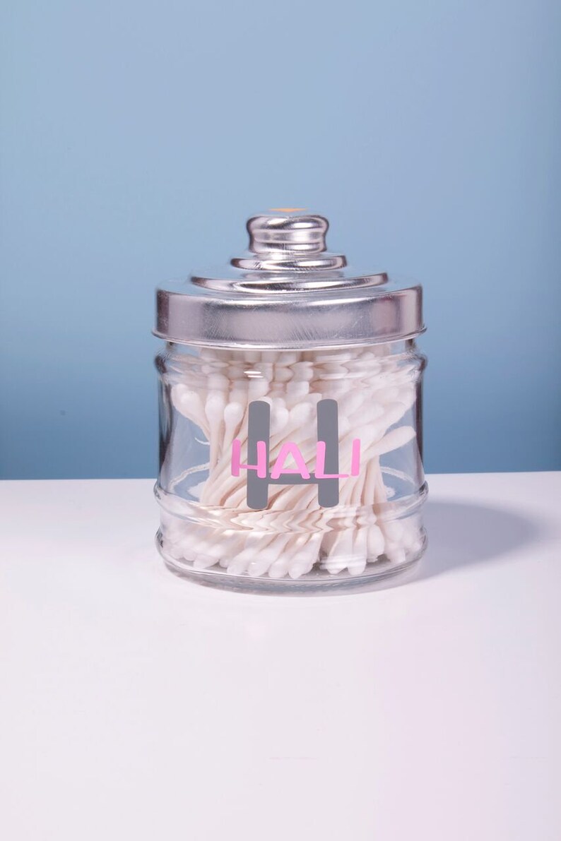 May include: A clear glass jar with a silver lid filled with cotton swabs. The jar has a gray label with the word "HALI" written in pink letters.