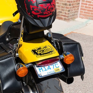 May include: A yellow motorcycle with black leather saddlebags and a black helmet with a red and black graphic. The motorcycle has a license plate that reads "M147C CYCLE JUN".