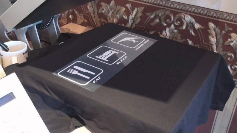 Puede incluir: Una camiseta negra con un dise&ntilde;o gr&aacute;fico blanco de tres iconos. Los iconos son un tenedor y un cuchillo, una cama y un ala delta. El texto debajo de los iconos dice "Comer, Dormir, Volar en ala delta"