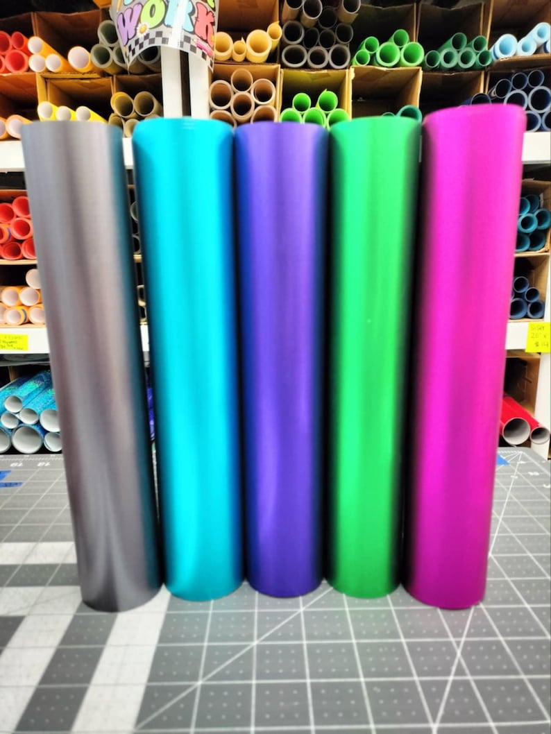 May include: Five rolls of metallic vinyl in different colors: gray, teal, purple, green, and pink. The rolls are arranged in a row on a gray grid surface.