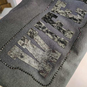 May include: Dark gray fabric with the word "LIFE" in a textured, patterned design. The word is outlined with small, black beads. The fabric appears to be a sweatshirt or similar garment.