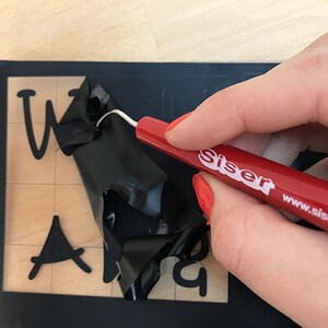 May include: A red weeding tool with the text "Siser www.siserna.com" is being used to remove excess vinyl from a black vinyl sheet with the words "Welcome" and "And" cut out.