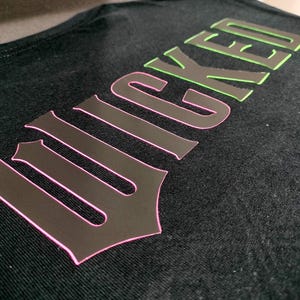 Może przedstawiać: Czarny T-shirt z nadrukiem słowa "Wicked" w stylu gotyckim z r&oacute;żowymi i zielonymi konturami.