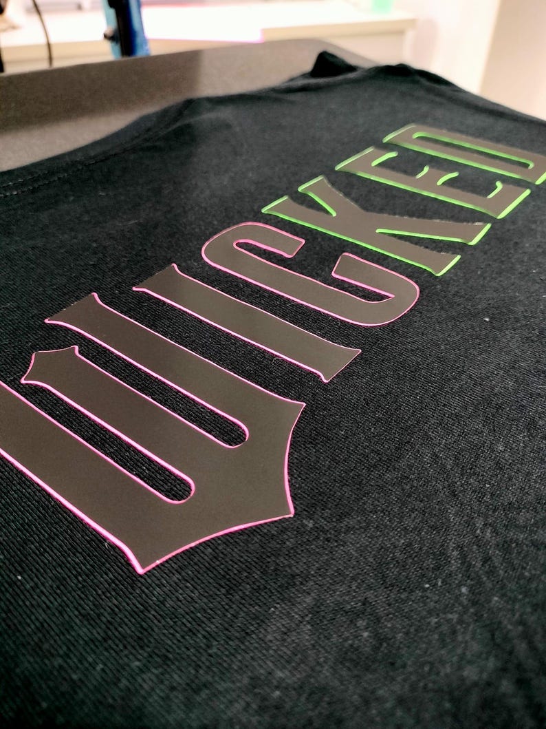 Może przedstawiać: Czarny T-shirt z nadrukiem słowa "Wicked" w stylu gotyckim z r&oacute;żowymi i zielonymi konturami.