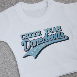 May include: White t-shirt with a blue and black graphic that says "Cheer Team Daredevils" with the text "Sister Dig Patterns" underneath.