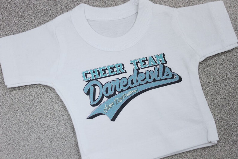 May include: White t-shirt with a blue and black graphic that says "Cheer Team Daredevils" with the text "Sister Dig Patterns" underneath.