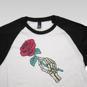 May include: A white and black baseball tee shirt featuring a red rose held by a skeletal hand. The rose has green leaves and a light blue stem.