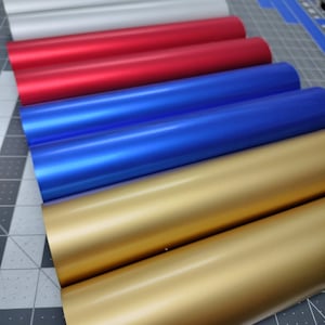 May include: A stack of five rolls of vinyl adhesive film in various colors, including silver, red, blue, and gold. The rolls are arranged in a graduated color pattern, with the silver roll at the top and the gold roll at the bottom.