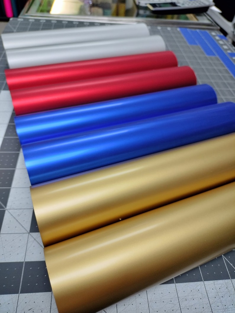May include: A stack of five rolls of vinyl adhesive film in various colors, including silver, red, blue, and gold. The rolls are arranged in a graduated color pattern, with the silver roll at the top and the gold roll at the bottom.