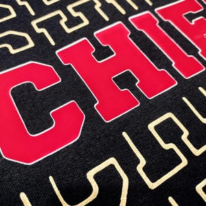 Puede incluir: Una sudadera negra con un gr&aacute;fico rojo de "CHIEFS" en la parte delantera. El fondo de la sudadera tiene un patr&oacute;n geom&eacute;trico dorado.