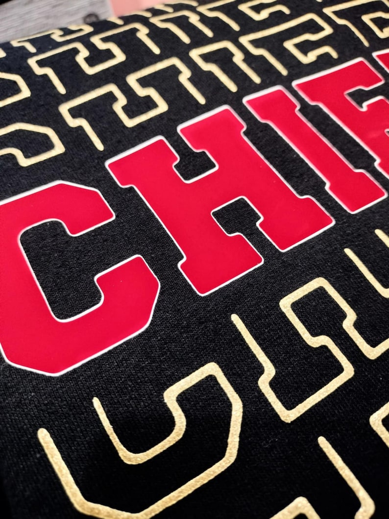 Puede incluir: Una sudadera negra con un gr&aacute;fico rojo de "CHIEFS" en la parte delantera. El fondo de la sudadera tiene un patr&oacute;n geom&eacute;trico dorado.