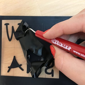 Puede incluir: Una herramienta de deshierbe roja con el texto "Siser www.siserna.com" se utiliza para eliminar el exceso de vinilo de una l&aacute;mina de vinilo negra con las palabras "Welcome" y "And" recortadas.