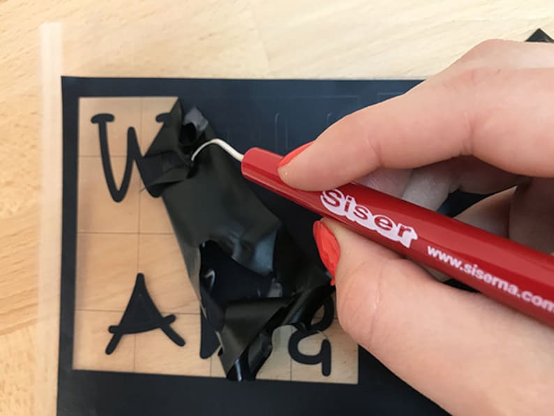 Puede incluir: Una herramienta de deshierbe roja con el texto "Siser www.siserna.com" se utiliza para eliminar el exceso de vinilo de una l&aacute;mina de vinilo negra con las palabras "Welcome" y "And" recortadas.