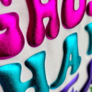 Op de afbeelding: Close-up van een witte stof met de woorden "YOU HAVE" geschreven in reliëf, glanzende letters. De letters zijn in een bubbelschrift en zijn roze en blauw.