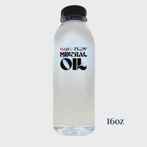 Puede incluir: Una botella de plástico transparente con tapa negra, llena de líquido transparente. La etiqueta dice "MAGIC FLOW MINERAL OIL" en texto colorido. La botella está etiquetada como "16oz".
