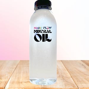 May include: A clear plastic bottle with a black cap, filled with a transparent liquid. The label reads "MAGIC FLOW MINERAL OIL" in a stylized font. The bottle sits on a wooden surface against a gradient pink and white background.