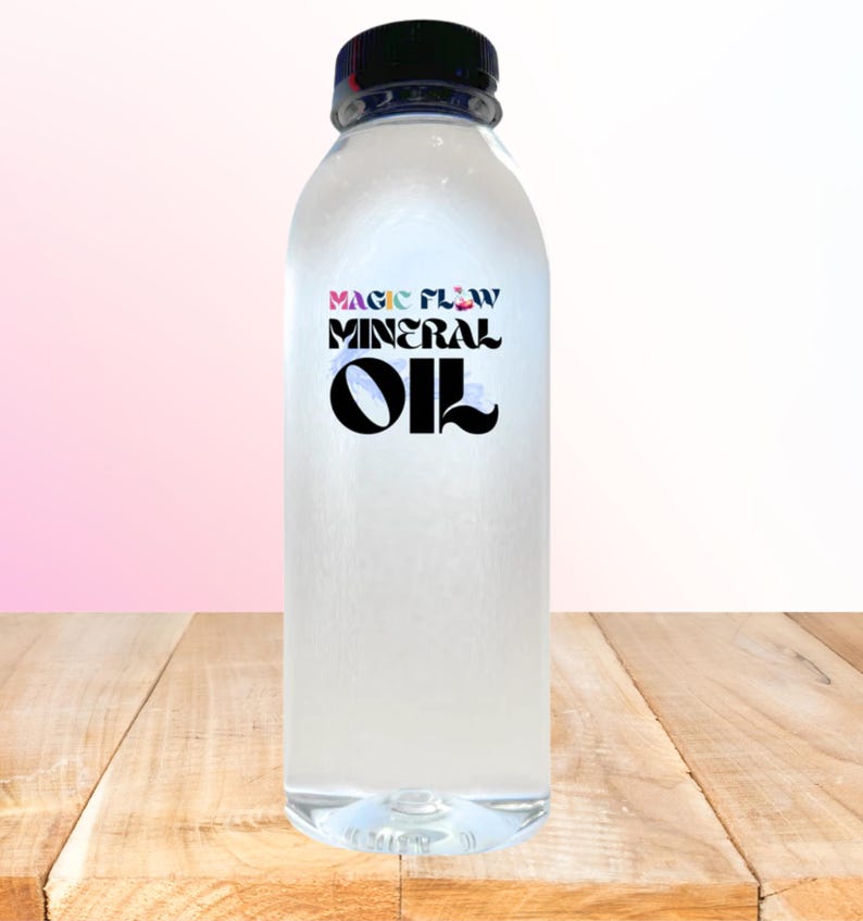 May include: A clear plastic bottle with a black cap, filled with a transparent liquid. The label reads "MAGIC FLOW MINERAL OIL" in a stylized font. The bottle sits on a wooden surface against a gradient pink and white background.