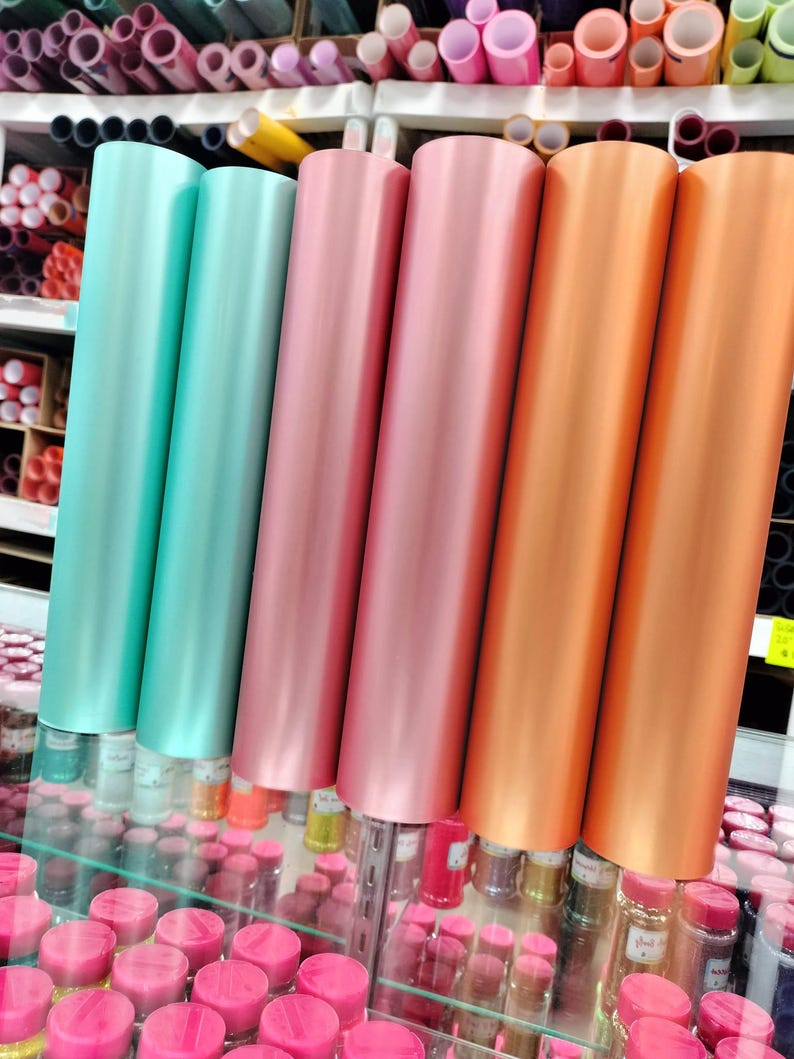 May include: Four rolls of metallic vinyl in different colors: teal, rose gold, copper, and orange. The rolls are stacked on a shelf in a store.