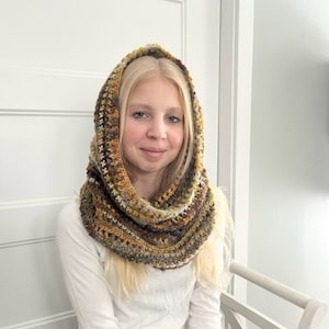May include: A woman wearing a brown, yellow, and white crocheted cowl with a hood. The cowl is made of a soft, textured yarn and has a loose, flowing design.