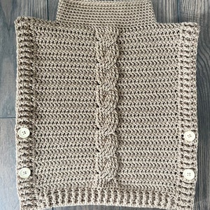 K&ouml;nnte beinhalten: Ein beigefarbener, handgeh&auml;kelter Cowl-Neck-W&auml;rmer mit einem Zopfmuster in der Mitte. Der Nackenw&auml;rmer hat einen hohen Kragen und wird mit zwei Kn&ouml;pfen auf jeder Seite befestigt. Der Artikel ist aus weichem, strukturiertem Garn gefertigt.