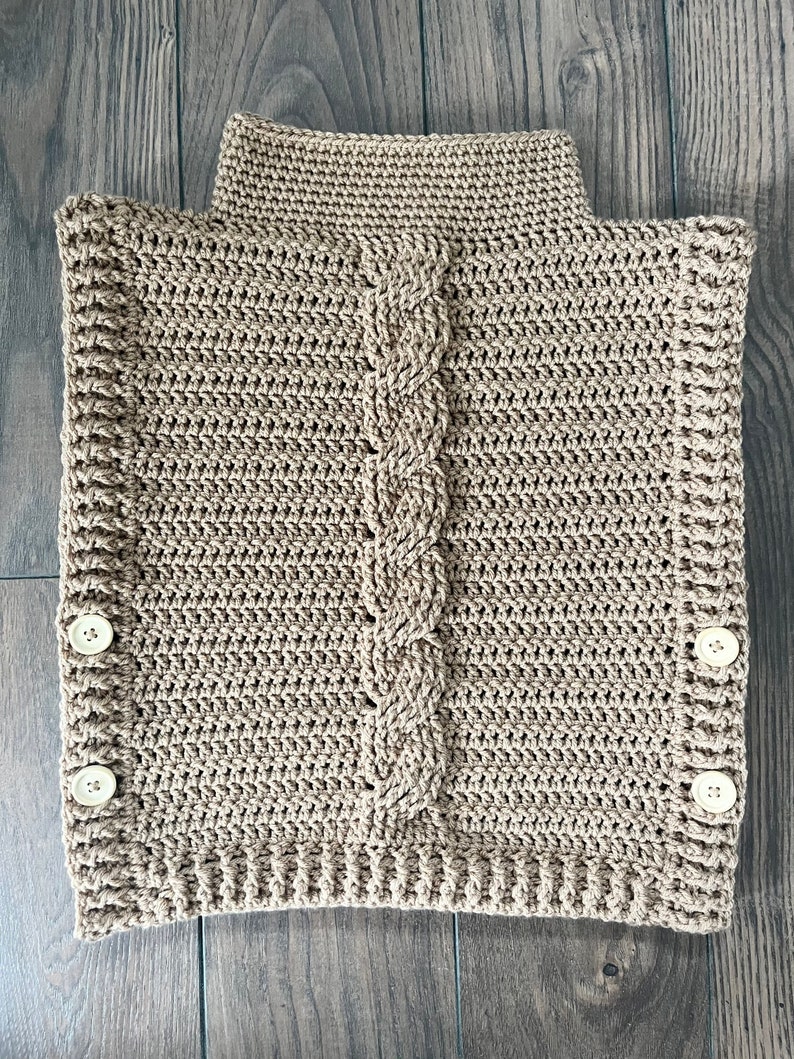 K&ouml;nnte beinhalten: Ein beigefarbener, handgeh&auml;kelter Cowl-Neck-W&auml;rmer mit einem Zopfmuster in der Mitte. Der Nackenw&auml;rmer hat einen hohen Kragen und wird mit zwei Kn&ouml;pfen auf jeder Seite befestigt. Der Artikel ist aus weichem, strukturiertem Garn gefertigt.