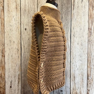 K&ouml;nnte beinhalten: Eine hellbraune H&auml;kelweste mit hohem Kragen und Knopfverschluss. Die Weste wird auf einer Schaufensterpuppe pr&auml;sentiert und weist ein strukturiertes Stichmuster auf. Die Kn&ouml;pfe sind hellbeige. Die Weste ist handgefertigt.