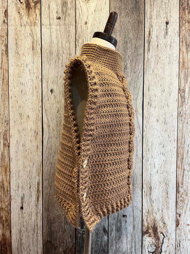 K&ouml;nnte beinhalten: Eine hellbraune H&auml;kelweste mit hohem Kragen und Knopfverschluss. Die Weste wird auf einer Schaufensterpuppe pr&auml;sentiert und weist ein strukturiertes Stichmuster auf. Die Kn&ouml;pfe sind hellbeige. Die Weste ist handgefertigt.